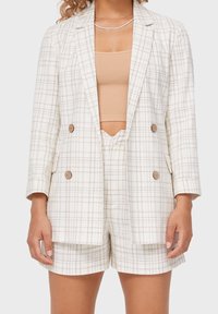 Blazer à carreaux beige clair et crème, présentant un design croisé avec des boutons en bois, accompagné d'un short assorti.