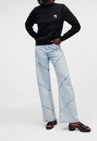 Czarna bluza z fakturowanej tkaniny i białym logo, zestawiona z jasnoniebieskimi dżinsami o szerokich nogawkach z przekątnym przeszyciem oraz czarnymi sandałami na słupku.