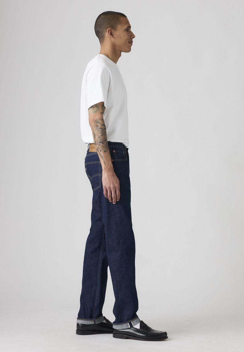 Joven con tatuajes de perfil, lleva una camiseta blanca, vaqueros azul oscuro con puños y zapatos de vestir negros sobre un fondo liso.