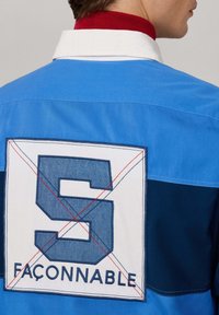Blauwe en marineblauwe katoenen overhemd met een witte achterpatch waarop een grote "S" en het woord "FAÇONNABLE" staat, omrand met contrasterende stitching.