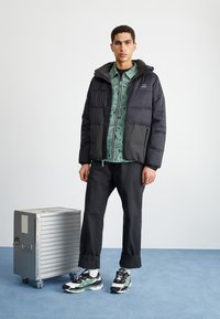 Black puffer jacket with hood, mint green patterned shirt beneath, dark trousers, and black and green sneakers beside a silver metallic suitcase.
