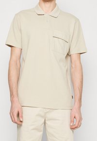 Polo beige avec un col classique, une patte de boutonnage à deux boutons et une poche à rabat. Le tissu est doux et lisse, avec des manches courtes.