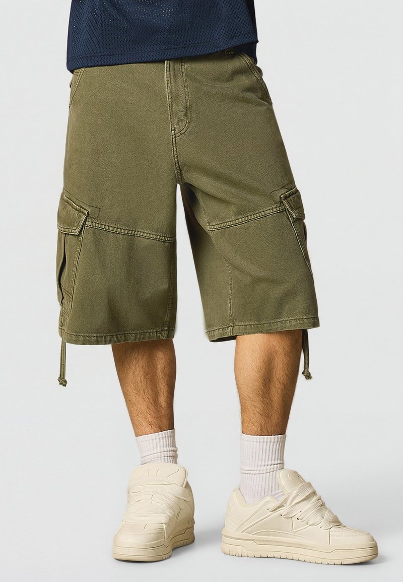 Man draagt olijfgroene cargoshorts met zijzakken, witte sportsokken en gebroken witte sneakers, staand tegen een effen achtergrond.