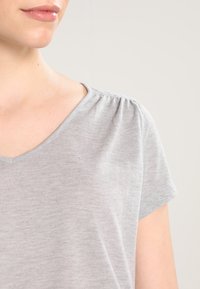 Šedý top s krátkým rukávem, který má výstřih do V a jemnou texturu s nabranými detaily na ramenou. Lehký a neformální design.