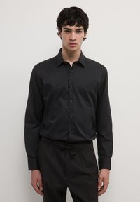 Junger Mann mit dunklem, lockigem Haar, der ein schwarzes Hemd mit Knöpfen und schwarze Jogginghosen trägt, steht vor einem schlichten, hellen Hintergrund.