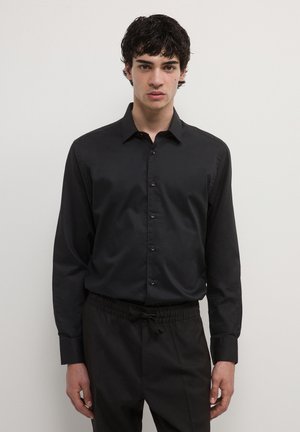 Young man with dark curly hair wearing a black button-up shirt and black drawstring pants, standing against a plain light background.