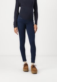 Jeans skinny bleu foncé à taille haute, fabriqués en tissu extensible et avec une texture lisse. Associés à des chaussures à lacets marron.
