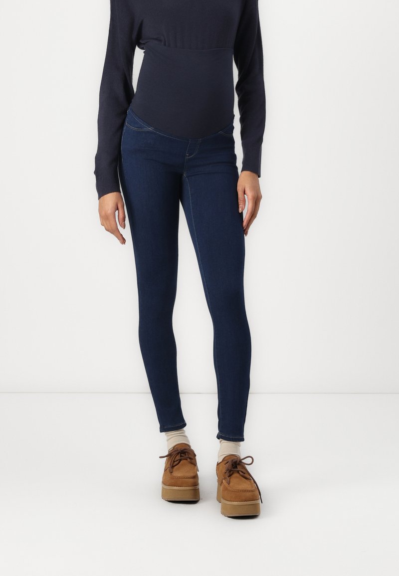 Jeans skinny bleu foncé à taille haute, fabriqués en tissu extensible et avec une texture lisse. Associés à des chaussures à lacets marron.