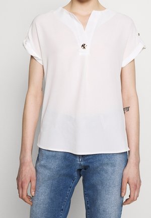 Blusa blanca de manga corta con escote en V y detalle de botones en el cuello. Tela ligera con una textura suave, combinada con jeans azules.