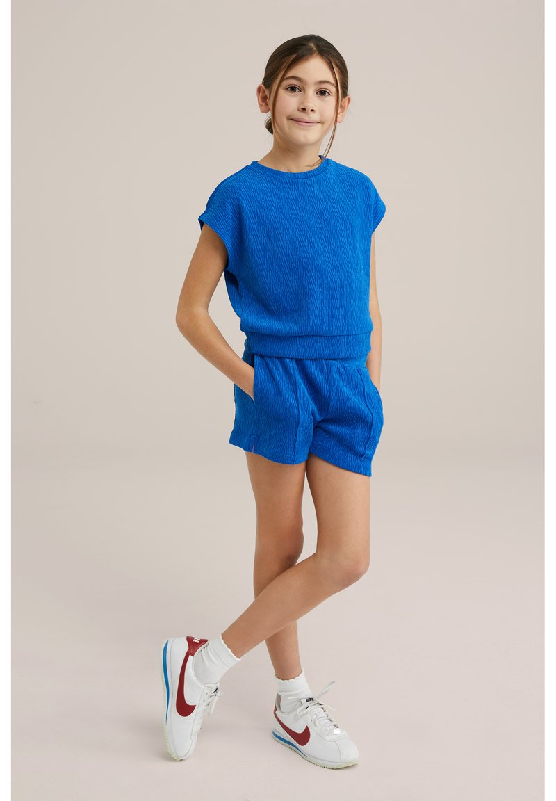 Modrý pletený top s texturou a odpovídající šortky. Oblečení se vyznačuje volným střihem, krátkými rukávy a kapsami, doplněným o bílé tenisky.