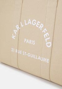 Sac fourre-tout en tissu beige avec un texte embouti blanc : "KARL LAGERFELD PARIS 21 RUE ST-GUILLAUME." Le texte suit la courbe de la couture supérieure. Texture lisse.