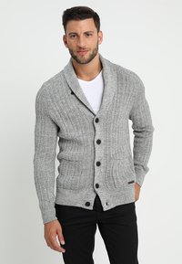 Grå strikket cardigan med sjalkrage, med vertikale ribbinger, fem brune knapper og to frontlommer. Mykt, teksturert stoff.