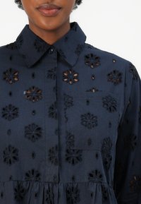 Camicia blu navy con ricami floreali a taglio, colletto, apertura frontale con bottoni e taschino sul petto. Presenta un tessuto testurizzato e un orlo a peplo.