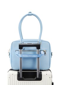 Bolso de mano de cuero azul claro con una textura suave, asas superiores y un bolsillo con cremallera en la parte frontal, reposando sobre una maleta blanca con ruedas.