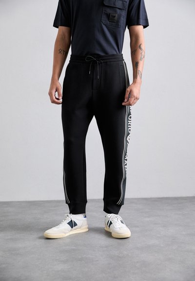 Homme portant un pantalon de jogging noir avec cordon de serrage et rayures latérales, et des sneakers blanches, se tenant sur un sol gris contre un mur uni.