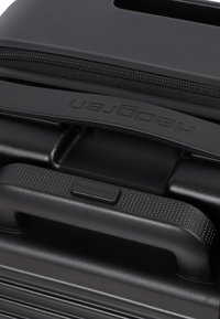 Gros plan d'une poignée de valise rigide noire avec une prise texturée et une fermeture éclair, estampillée du nom Hedgren.