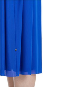 Blå klänning i lätt tyg, med en flödande design, veck och en liten metallaccent vid fållen.