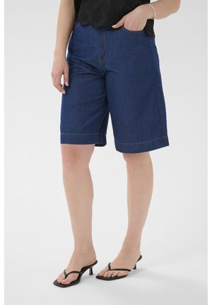 Kobieta ubrana w ciemnoniebieskie szorty dżinsowe do kolan, czarny bezrękawnik i czarne sandały na obcasie, stoi z jedną ręką w kieszeni.