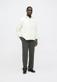 Világos színű, gombos dzseki puha anyagból, gallérral, sötét, egyenes szárú nadrággal és fényes barna cipővel párosítva.