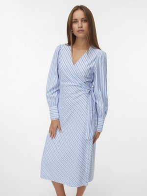 Νεαρή γυναίκα με φόρεμα τύπου wrap σε γαλάζια και λευκή ρίγα, με μακριά φουσκωτά μανίκια και δεμένο φιόγκο στη μέση, στέκεται μπροστά σε απλό φόντο.