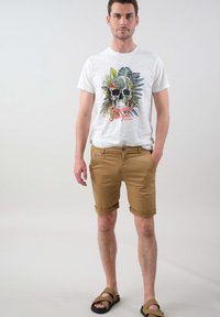 T-shirt blanche avec un graphisme coloré de crâne et de feuillage, associée à un short beige. Le short est retroussé à l'ourlet. Des tongs complètent le look.