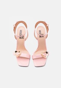 Ροζ πέδιλα ψηλά τακούνια από δερματίνη με λουράκια στον αστράγαλο και χρυσά αγκράφες σε σχήμα καρδιάς, με την επωνυμία "Love Moschino" στην εσωτερική σόλα.