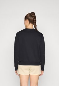 Sweatshirt preto com gola redonda e mangas longas. Apresenta uma bainha e punhos canelados. Usado com calções bege. Textura suave.