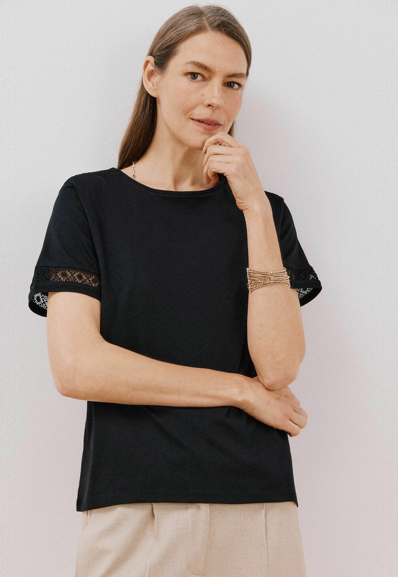 Sort kortærmet T-shirt med blonder på ærmerne, rund hals og en afslappet pasform, parret med beige bukser.