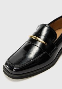 Black patent leather loafer with a flat sole, featuring a gold metallic accent across the strap and a smooth, glossy finish.