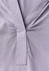 Tessuto lavanda con scollo a V caratterizzato da una piega centrale e una toppa rettangolare alla base dello scollo. Texture liscia.