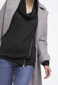 Grau langer Mantel über einem schwarzen Oberteil mit Kapuze und seitlichem Reißverschluss, kombiniert mit blauen Jeans. Der Stoff hat einen leichten Schimmereffekt.