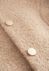 Tessuto beige con trama a rilievo, caratterizzato da fibre ad anello e bottoni metallici dorati. Il materiale presenta un aspetto morbido e peloso con dettagli argentati.