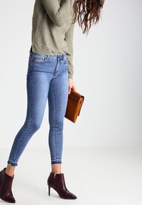 Světle olivový svetr s texturou, modré skinny džíny s roztrhanými okraji a vínové kotníkové boty. Drží hnědou koženou kabelku.