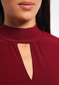 Burgunderfärgad blus med hög hals och en nyckelhålsöppning, med mjukt, slätt tyg och rynkdekor kring halsringningen.