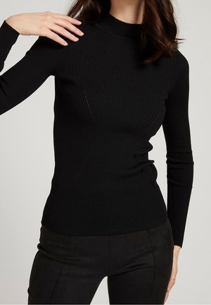 Kobieta w dopasowanym, czarnym, prążkowanym swetrze z długim rękawem i czarnych spodniach, pozująca z jedną ręką przy szyi, a drugą przy boku.