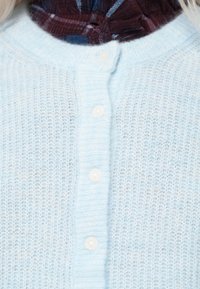 Zbliżenie na jasnoniebieski sweter dzianinowy z białymi guzikami na tle bordowej koszuli w wzory.