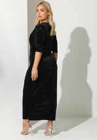 Blond kvinna i svart sammetsklänning i ankel-längd, bärande silver höga klackar och turkosa örhängen, ser över axeln.