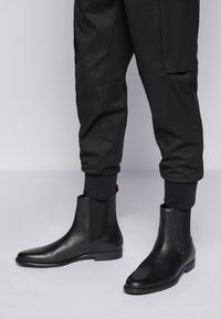 Bottes cheville en cuir noir avec panneaux élastiques sur les côtés, bout pointu et texture lisse. Portées avec un pantalon noir fuselé et des bordures côtelées.