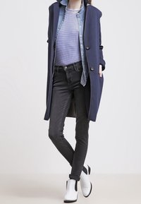 Marinblå lång kappa över en denimskjorta och en randig topp, kombinerad med svarta slim-fit jeans och vita ankelstövlar med svarta accenter.