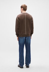 Brun sweatshirt med rundad halsringning, med kontrasterande ljus sömnad längs sömmarna, i kombination med lösa blå denimjeans.