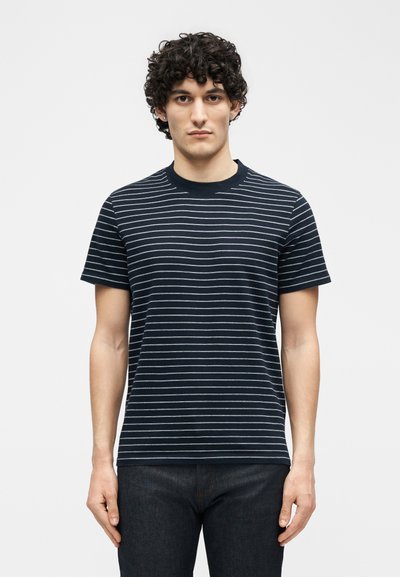 Νέος άνδρας με σγουρά μαλλιά που φοράει μπλε σκούρο μπλουζάκι με λεπτές λευκές οριζόντιες ρίγες, στέκεται μπροστά σε απλό λευκό φόντο.