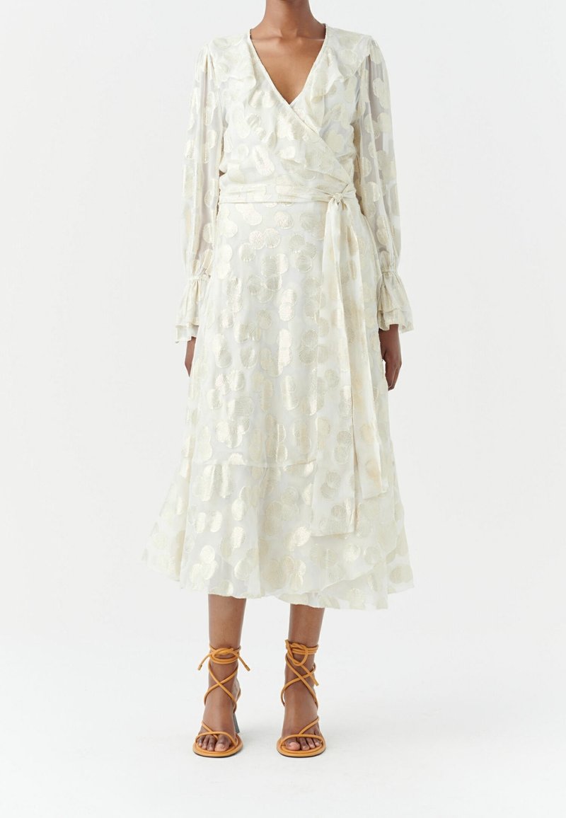 Kevyt kermaisen värinen kietaisumekko, jossa on kultaisia ympyräkuvioita. Ominaisuuksiin kuuluvat pitkät hihat, kerroksellinen hame ja yhteensopiva vyö. Yhdistettynä ruskeisiin sandaaleihin.