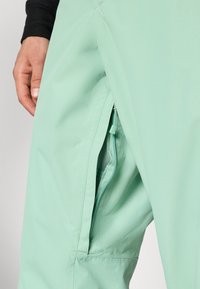 Pantaloni da esterno verde menta con zip laterale e fodera in rete. Tessuto liscio e leggero, progettato per comfort e funzionalità.