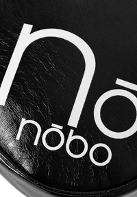 Ein schwarzes strukturiertes Material mit fettem weißen Text "nōbō" und einem kreisförmigen Design. Akzente umfassen eine glänzende Oberfläche und leichte Gebrauchsspuren.
