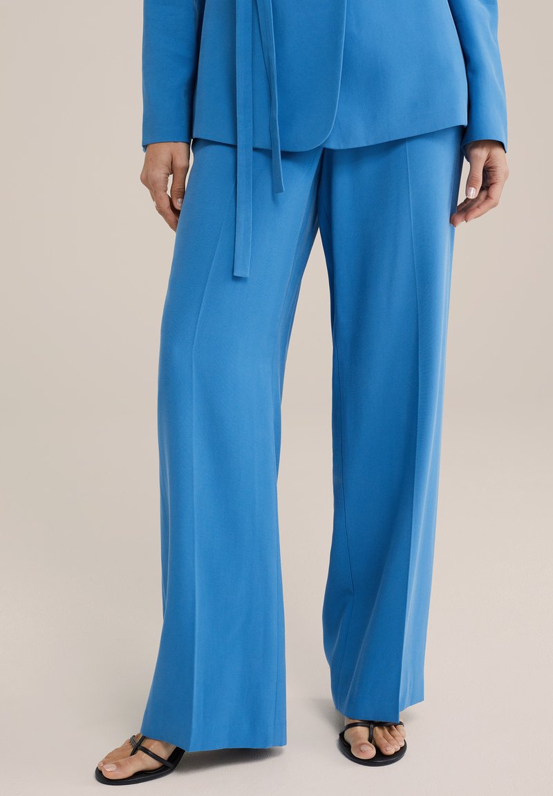 Modrý dvoudílný outfit obsahující podlouhý rukáv s uzlem a kalhoty s širokým střihem. Hladká textura, tmavé sandály s minimem popruhů.