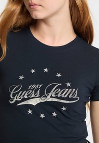Tamsiai mėlynas medvilninis marškinėliai su sidabriniu grafikiniu spaudiniu "Guess Jeans" ir 1981 metų numeriu, apipavidalintis mažomis žvaigždutėmis.