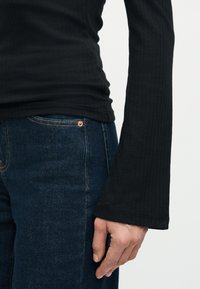 Δεξιά πλευρά ατόμου που φορά μαύρο ριμπ πλεκτό μακρυ μάνικο τοπ με φαρδύ μανίκι και σκούρο μπλε τζιν, με το χέρι χαλαρό στο πλάι.