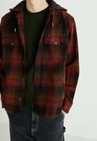 Camicia di flanella a quadri rossi e neri con cappuccio, dotata di chiusure a bottoni, due tasche sul petto e un tessuto testurizzato. Indossata sopra una maglietta verde.