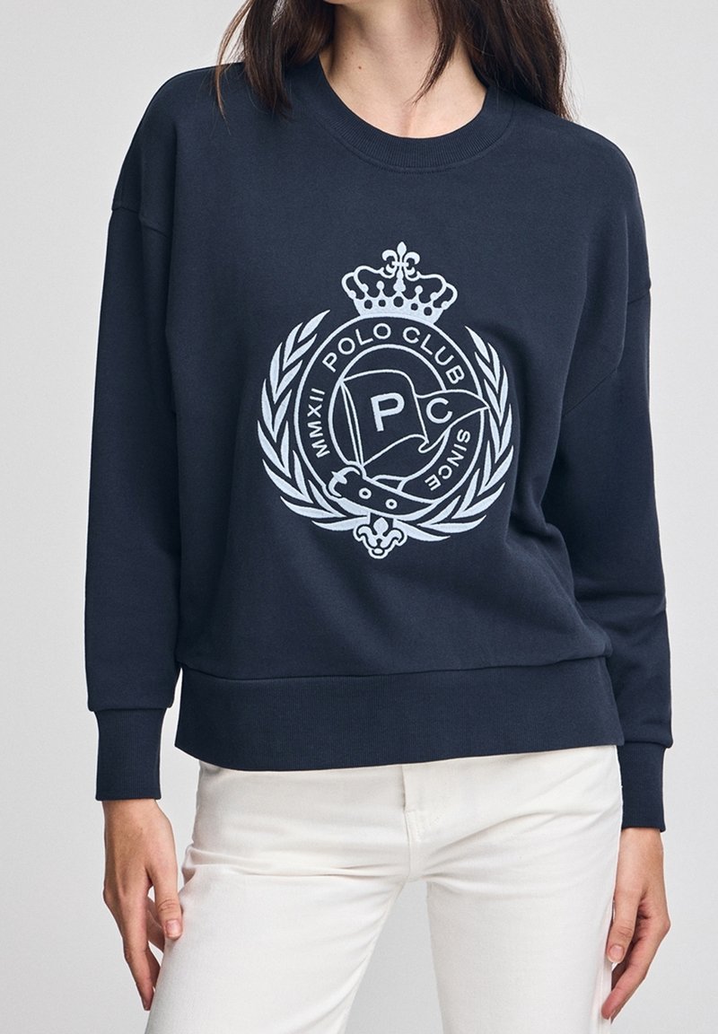 Femme portant un sweat-shirt bleu marine avec un écusson blanc Polo Club et un motif de laurier, associée à un pantalon blanc, debout devant un fond uni.