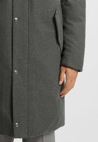 Grau melierter Wollmix-Mantel mit Knopfverschluss, seitlichen Taschen und geradem Schnitt. Der Stoff hat eine glatte, weiche Textur. Sauberes Design.